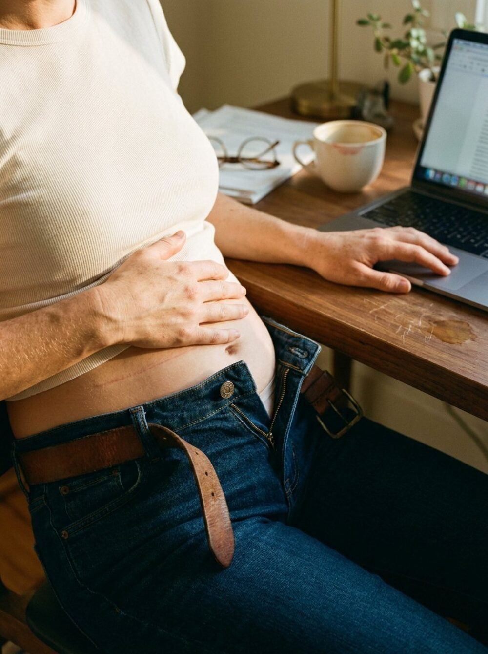 Cropped view of a woman seated at a wooden desk in late afternoon light. Her hand rests protectively on her bare stomach, her jeans are visibly unbuttoned with the waistband gaping open, and her other hand is on a laptop trackpad. The shot captures the unconscious daily moment of bloat shame that millions of women in their 30s experience without naming.